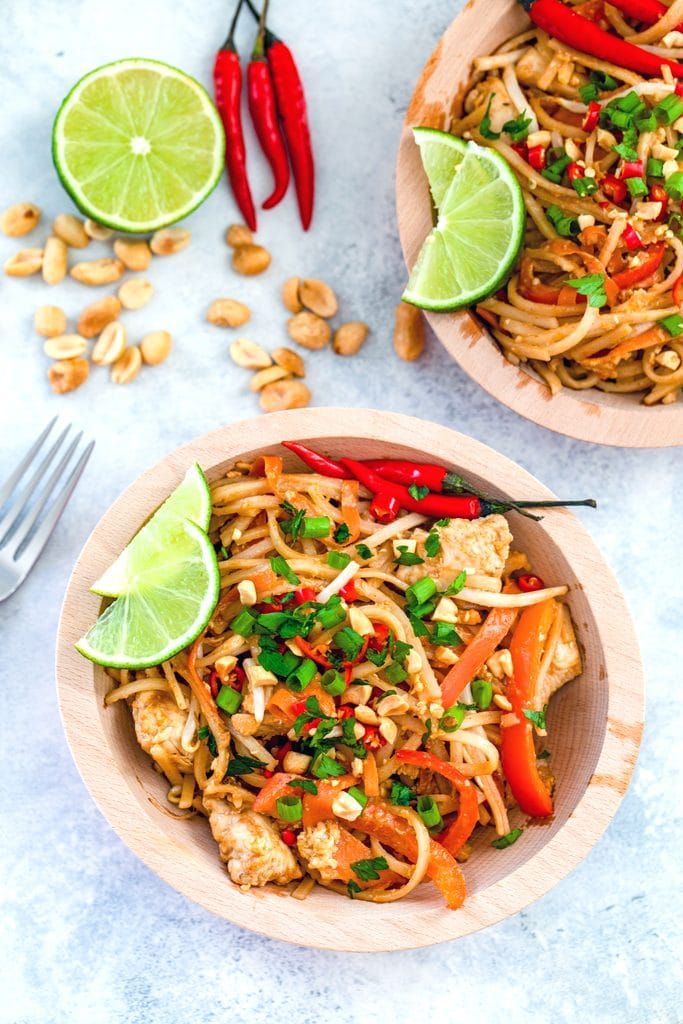 Direct overhead view of wooden bowl of chicken Pad Thai with peanuts, lime wedge, and red Thai chili peppers in the background, along with a second bowl of Pad Thai