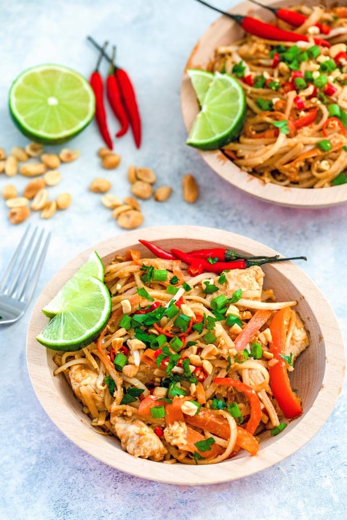 Overhead view of a bowl of Chicken pad Thai topped with scallions red chili peppers, peanuts, and limes, with peanuts, limes, peppers, and a second bowl of Pad Thai in the background