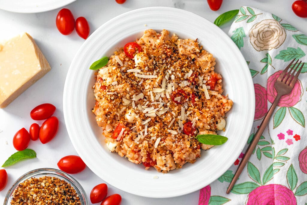 Landscape overhead view of a bowl of chicken parmesan risotto with cherry tomatoes, hunk of parmesan cheese, basil, and small bowl of panko in background