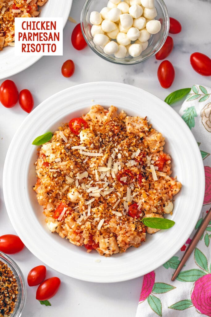 Overhead view of a bowl of chicken parmesan risotto topped with crispy panko with cherry tomatoes and mozzarella balls all round and recipe title at top