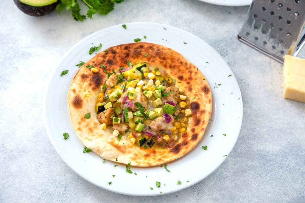 Landscape view of white plate with a chicken and summer vegetable tostada on it topped with avocado and cheese with cheese grater and block of cheese in background