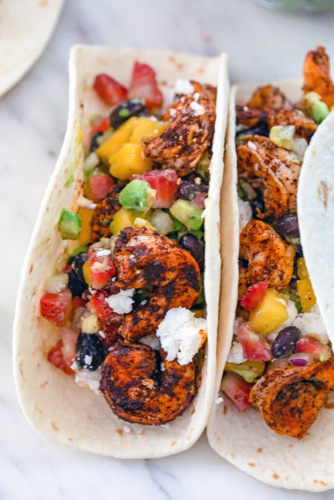 Close-up head-on view of two chili lime shrimp tacos with strawberry, mango, and avocado salsa and feta on top.