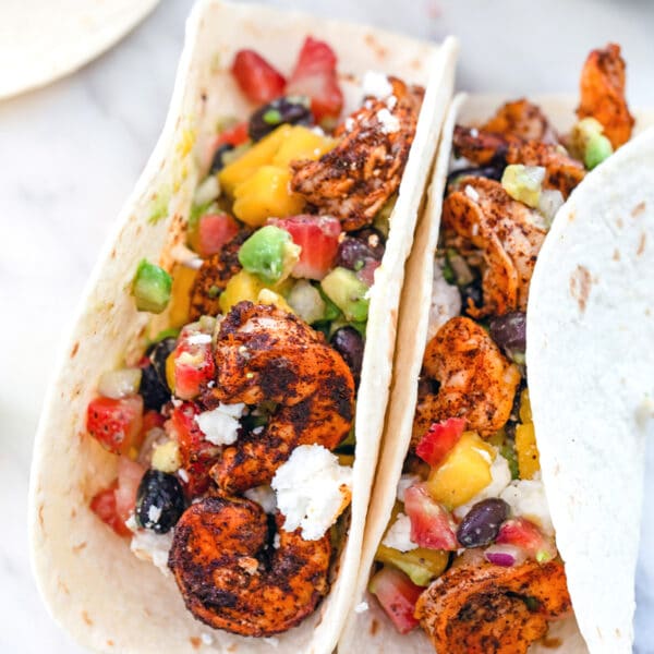 Overhead closeup view of two chili lime shrimp tacos with a strawberry, mango, avocado salsa and feta on a marble surface.