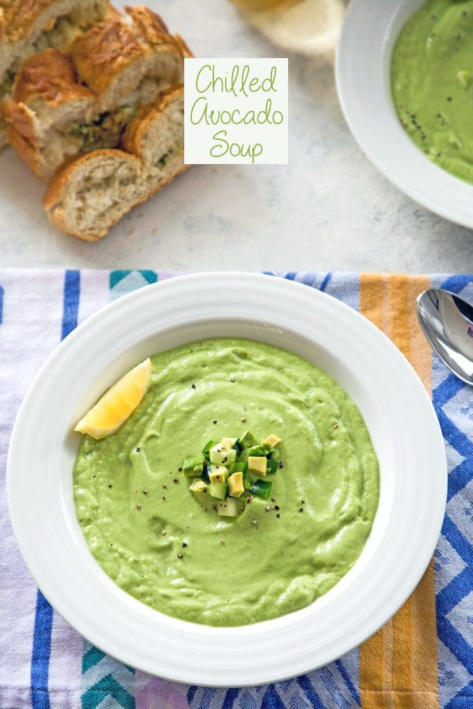 Overhead view of cold avocado soup in a white bowl on a colorful towel with loaf of sliced bread and second bowl in the background and "Chilled Avocado Soup" text at top