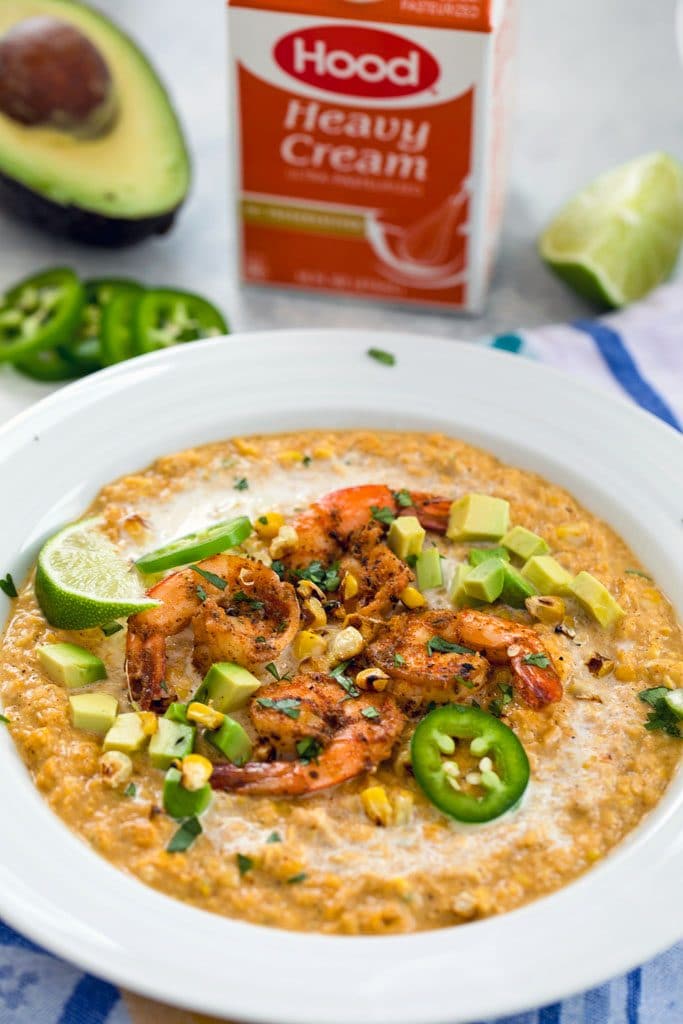 Head-on vide of chilled Mexican corn bisque topped with spicy shrimp, jalapeño slices, grilled corn kernels, diced avocado, and lime wedges with half an avocado, jalapeño slices and container of heavy cream in the background