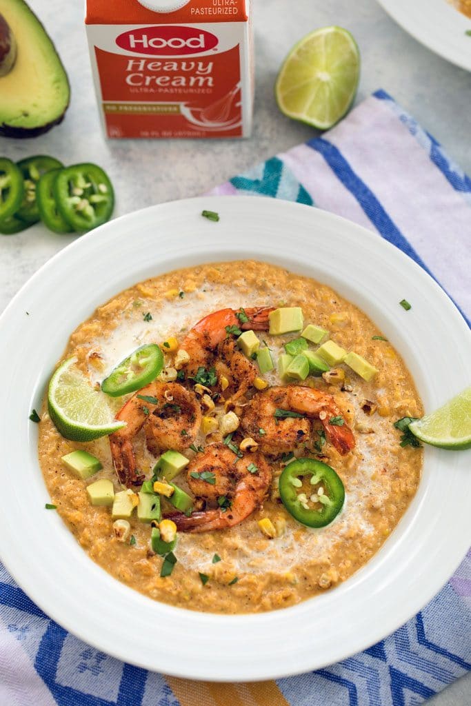 Overhead view of chilled Mexican corn bisque topped with shrimp, diced avocado, grilled corn kernels, sliced jalapeño, and lime wedges with heavy cream container avocado, lime, and jalapeños in the background