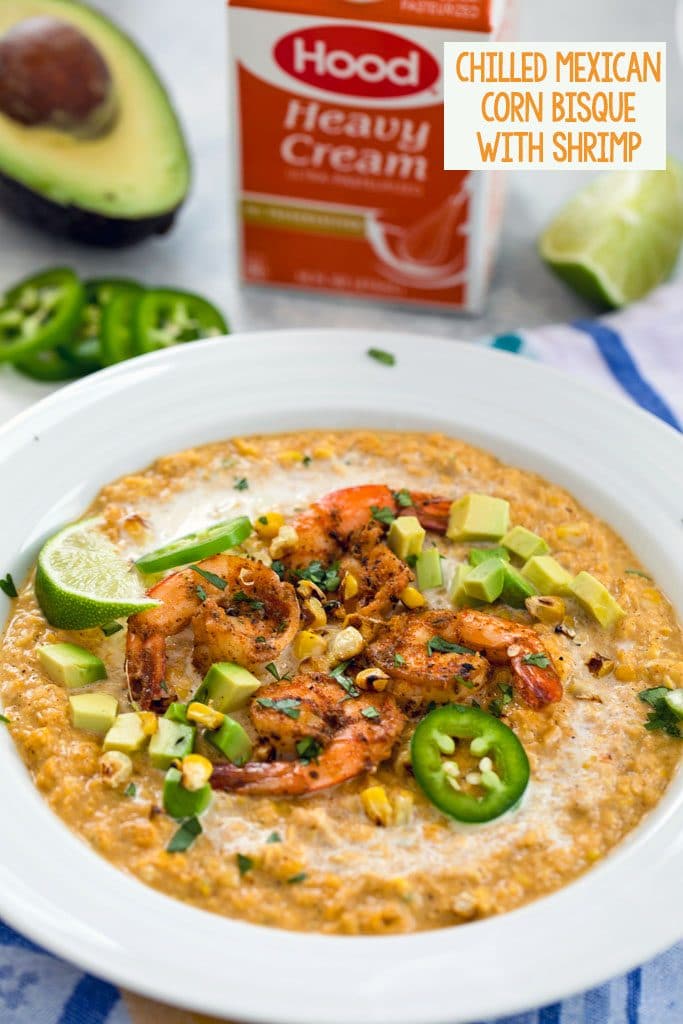 Head-on vide of chilled Mexican corn bisque topped with spicy shrimp, jalapeño slices, grilled corn kernels, diced avocado, and lime wedges with half an avocado, jalapeño slices and container of heavy cream in the background and "Chilled Mexican Corn Bisque with Shrimp" in text at the top