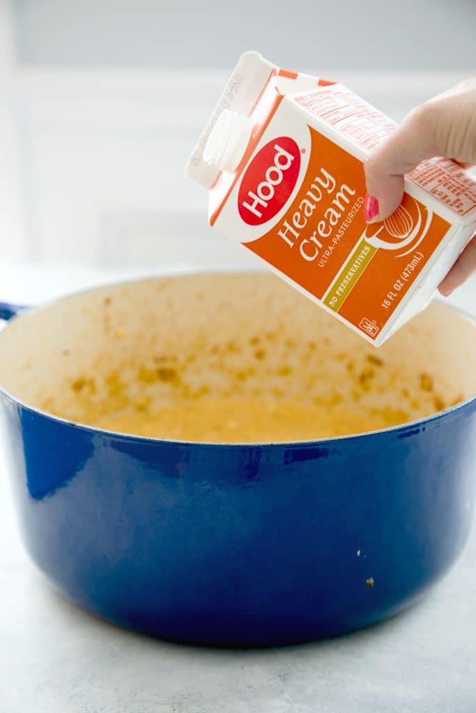 Blue dutch oven containing corn bisque with container of heavy cream being held above it ready to be poured