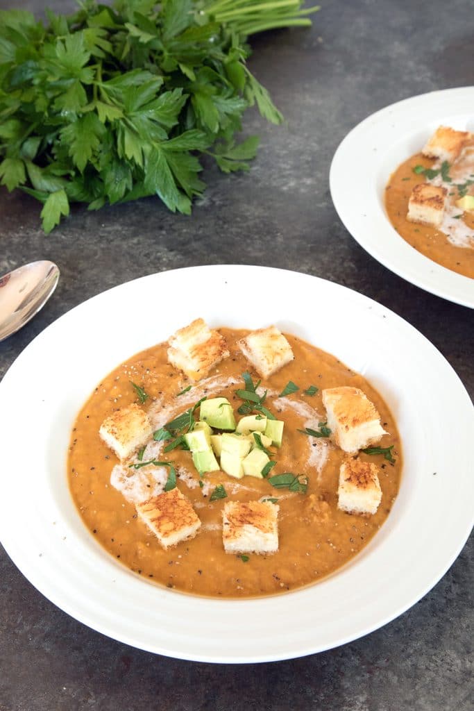 Head on view of a white bowl filled with butternut squash soup with grilled cheese croutons, diced avocado, and parsley garnishes, with bunch or parsley and spoon in the background