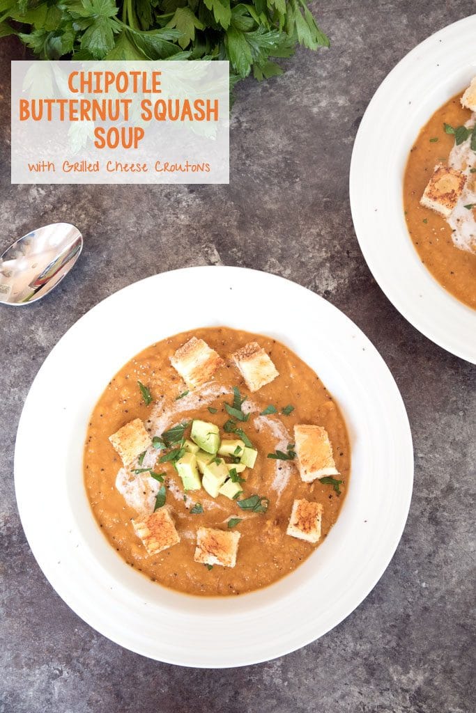 Overhead view of chipotle butternut squash soup in a white bowl with grilled cheese croutons and avocado garnish, parsley and a spoon in the background and recipe title at the top