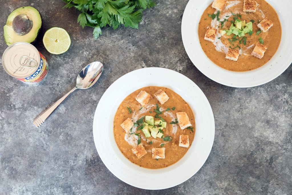 Landscape overhead view of two white bowls of chipotle butternut squash soup with spoon, can of chipotles, half a lime, half an avocado, and a bunch of parsley in the background