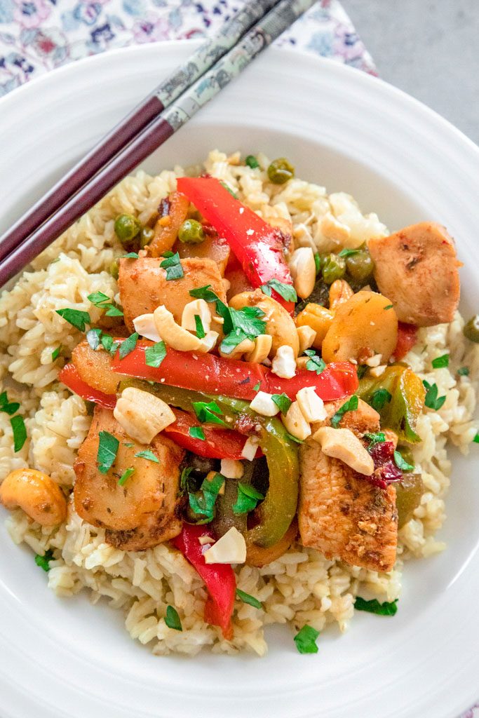 Overhead closeup view of chipotle chicken with cashews, red and green peppers, and peas over rice in a white bowl with chopsticks