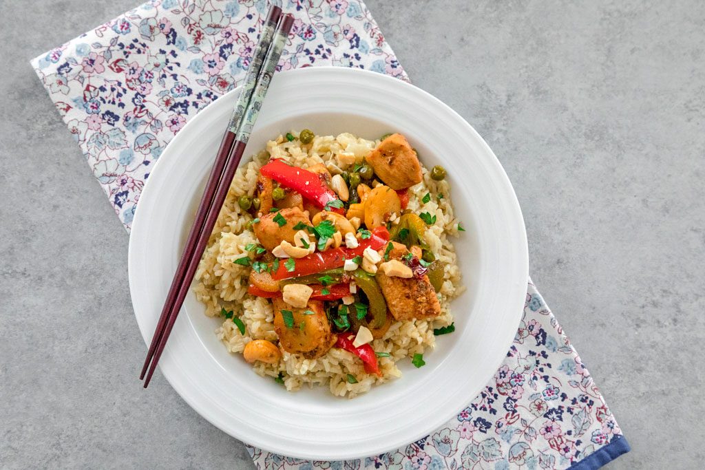 Landscape bird's eye view of white bowl filled with rice and chipotle chicken with cashews, red and green peppers, and peas with chopsticks resting on bowl