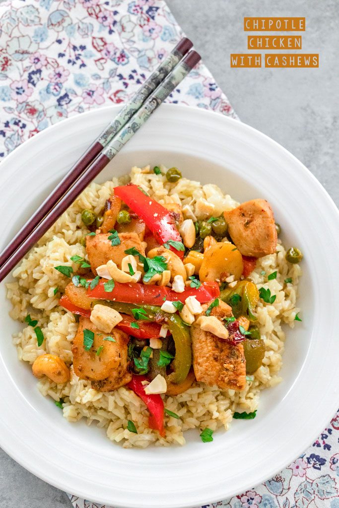 Overhead view of chipotle chicken with cashews, red and green peppers, and pea over rice in a white bowl with chopsticks and the recipe title at top