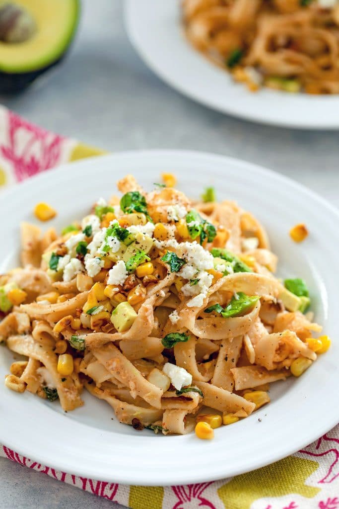 Close-up image of chipotle corn fettuccine on a white plate and green and pink napkin with avocado half and second plate in the background