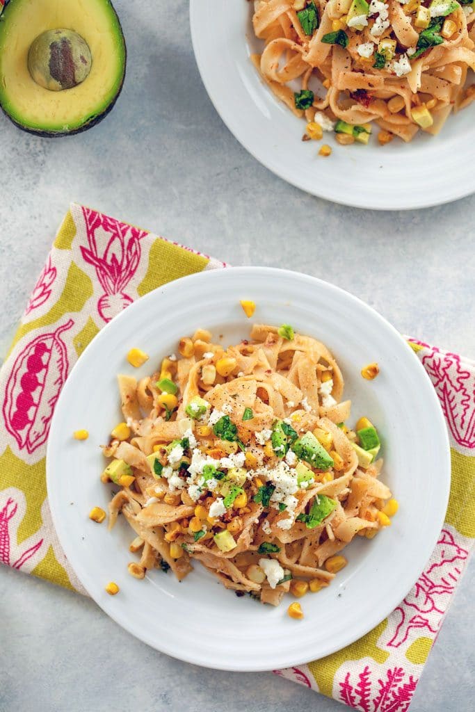 Bird's eye view of chipotle corn fettuccine on white plate and green and pink napkin with avocado half and second plate in the background