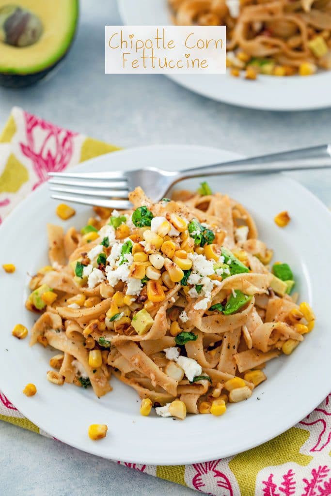 Head-on view of chipotle corn fettuccine on a white plate and green and pink towel with a fork, avocado half, and second plate in the background and "Chipotle Corn Fettuccine" text at top