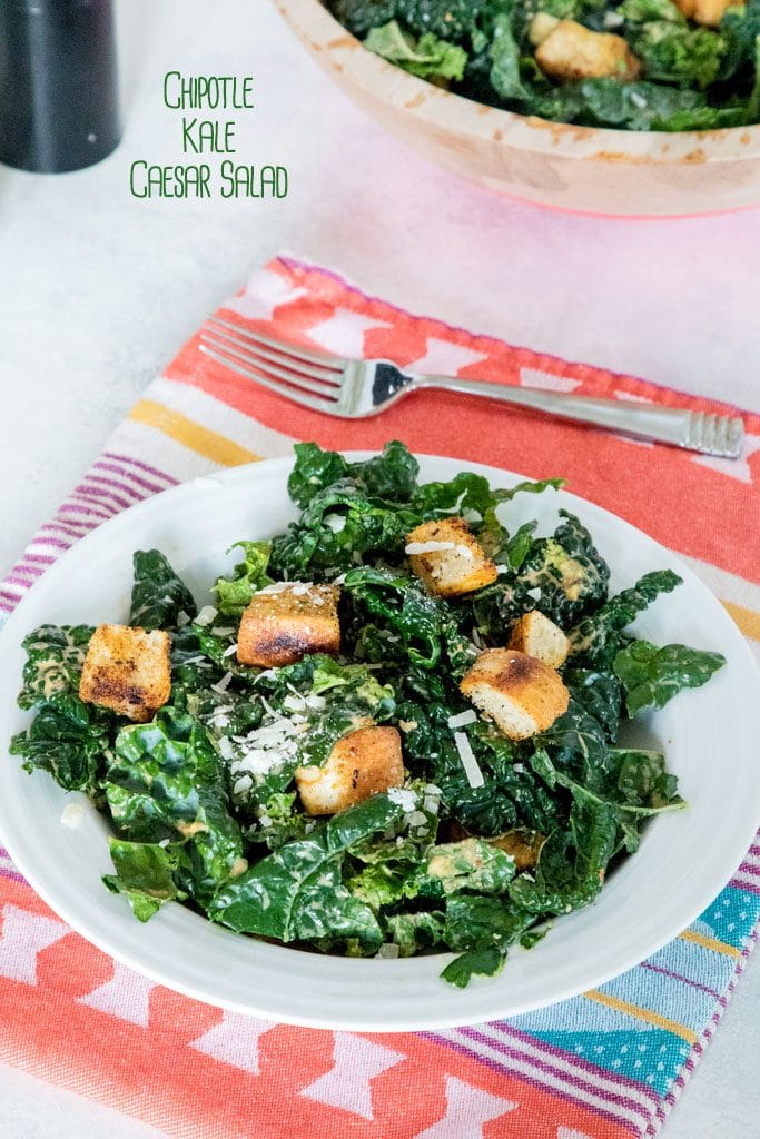 Chipotle kale Caesar salad in a white bowl on a red and yellow placemat and white surface with fork and large bowl of salad in the background and "Chipotle Kale Caesar Salad" text