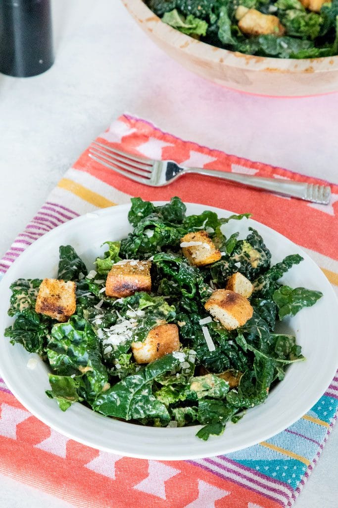 Chipotle kale Caesar salad in a white bowl on a red and yellow placemat and white surface with fork and large bowl of salad in the background