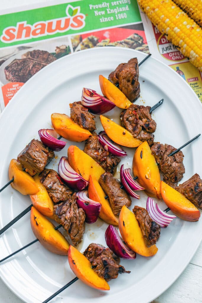 Overhead view of marinated steak, sliced peaches, and chopped onion on kabobs on a platter with grilled corn in the background