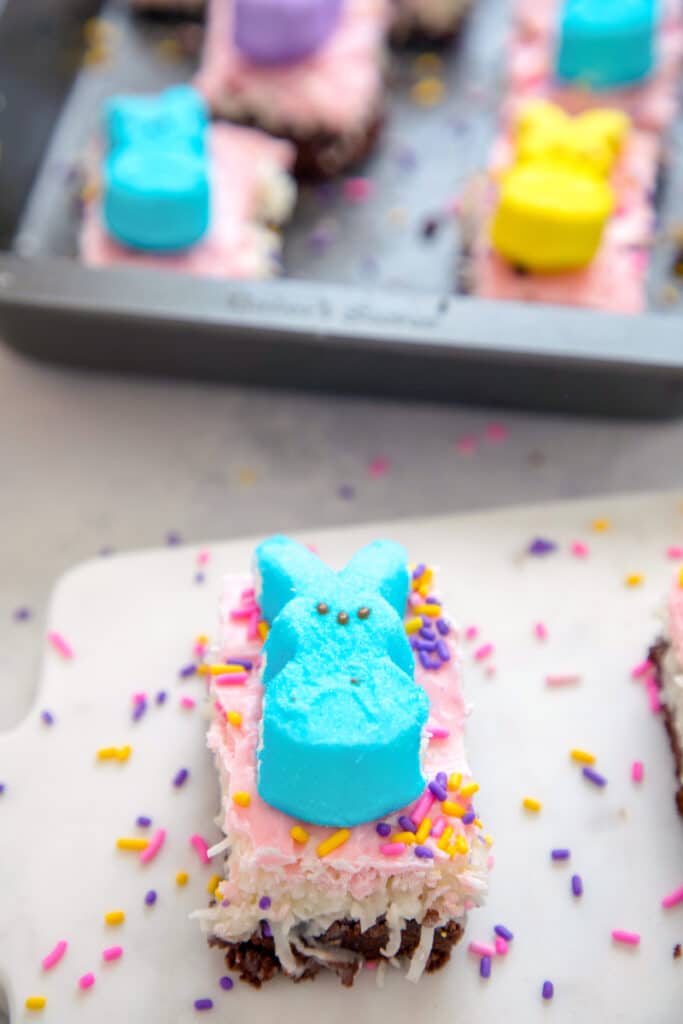 Overhead view of a chocolate coconut brownie bar with pink icing and a blue Peep on top with lots of sprinkles and a pan of more Peeps bars in the background