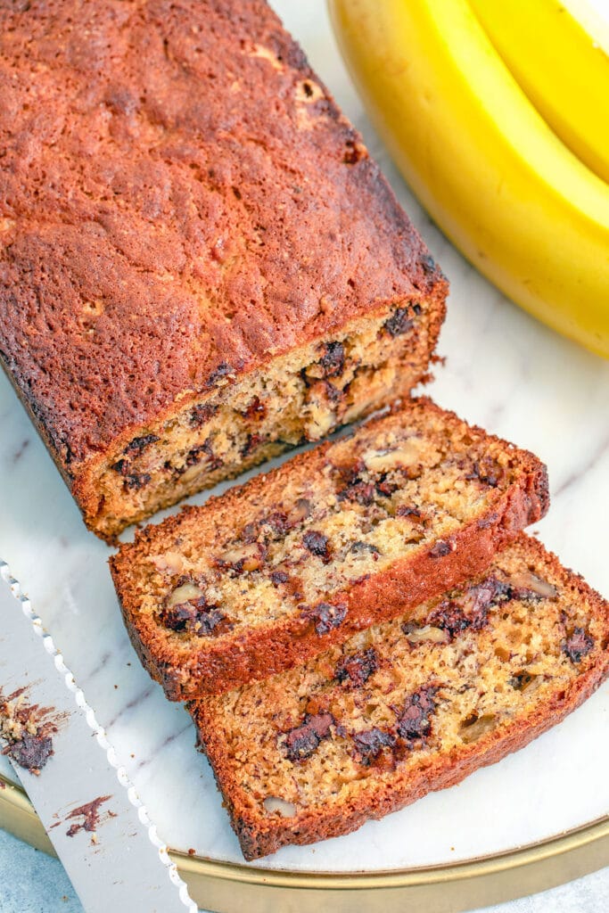Overhead closeup view of a loaf of chocolate chip banana bread with a couple slices cut a knife and bananas in the background.