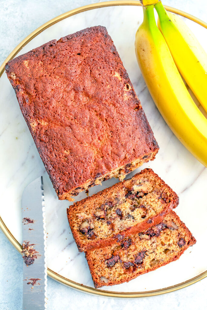 Bird's eye view of a loaf of chocolate chip banana bread with slices cut out on a marble tray with knife and bananas in background.