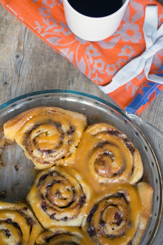 Overhead view of a dish of chocolate chip cinnamon rolls with butterscotch icing drizzled over the top and a cup of coffee on a tea towel in the background