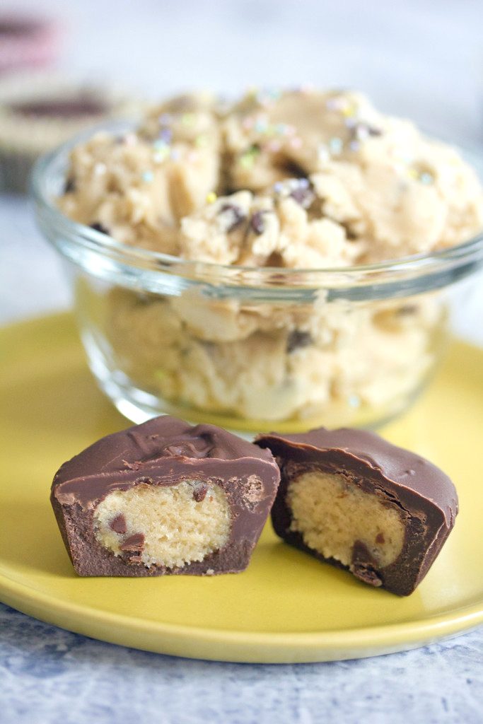 Head-on view of a chocolate-covered chocolate chip cookie dough cup cut in half on a yellow plate with bowl of cookie dough with sprinkles in the background