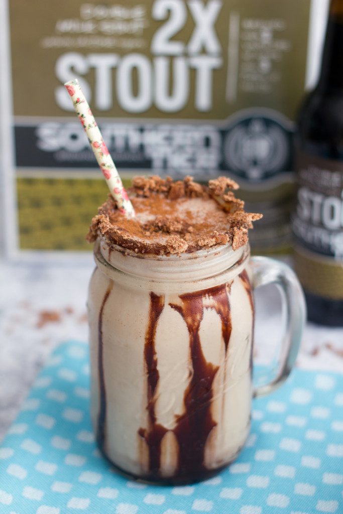 Head-on view of a Chocolate Cinnamon Toast Crunch stout  milkshake in a mason jar with chocolate syrup drizzle, cereal rim, and straw with beer in the background
