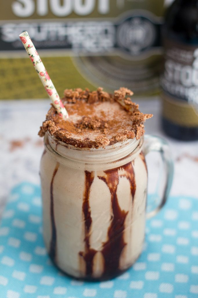 Head-on close-up view of a Chocolate Cinnamon Toast Crunch stout  milkshake in a mason jar with chocolate syrup drizzle, cereal rim, and straw with beer in the background