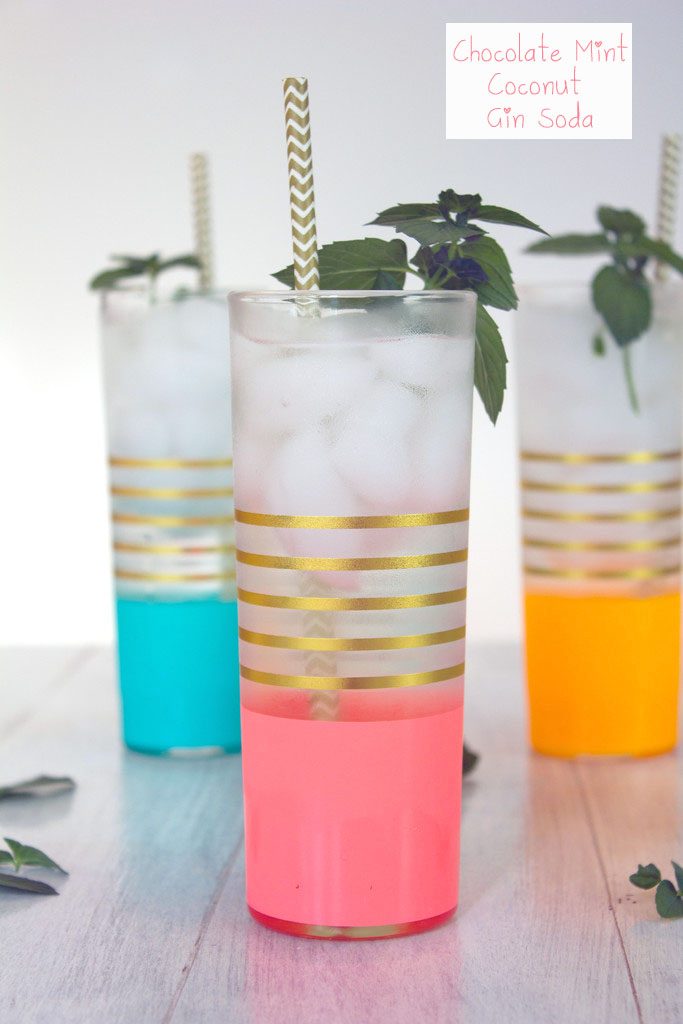 Head-on view of a pink and gold glass filled with chocolate mint coconut gin soda with gold and white straw and fresh chocolate mint with more cocktails in the background and recipe title at top