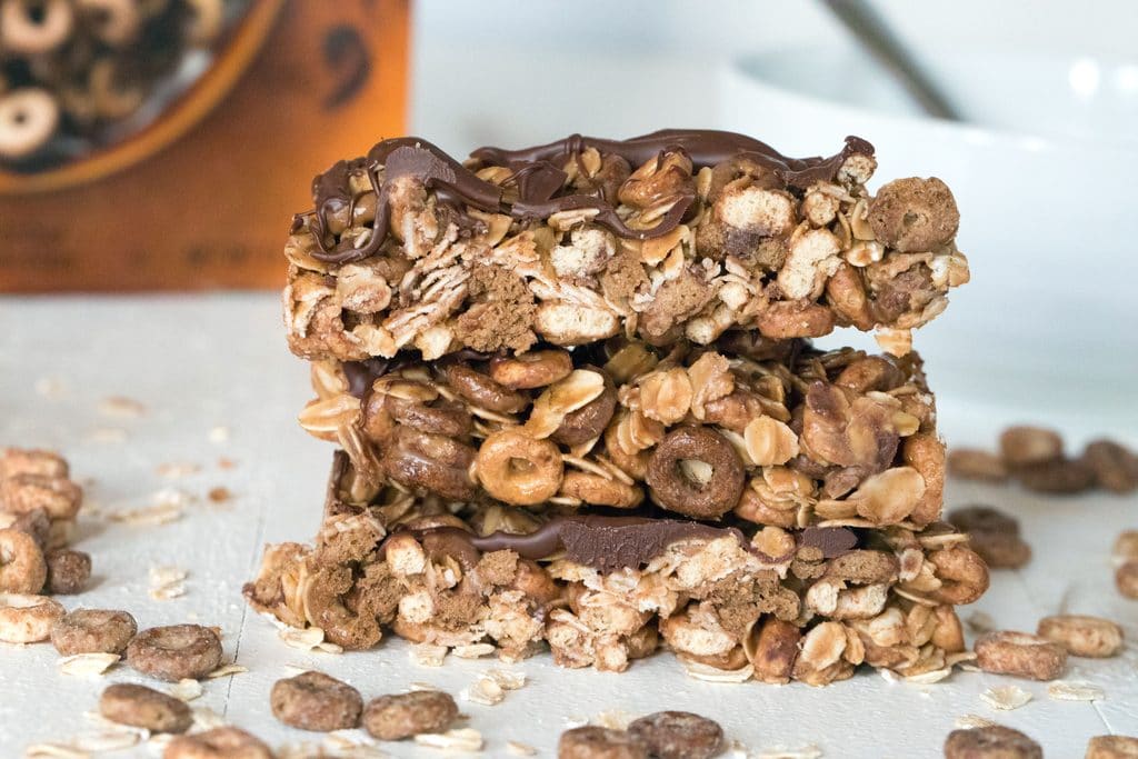 Landscape view of three chocolate peanut butter cheerios granola bars with cereal and oats around and cereal box in the background