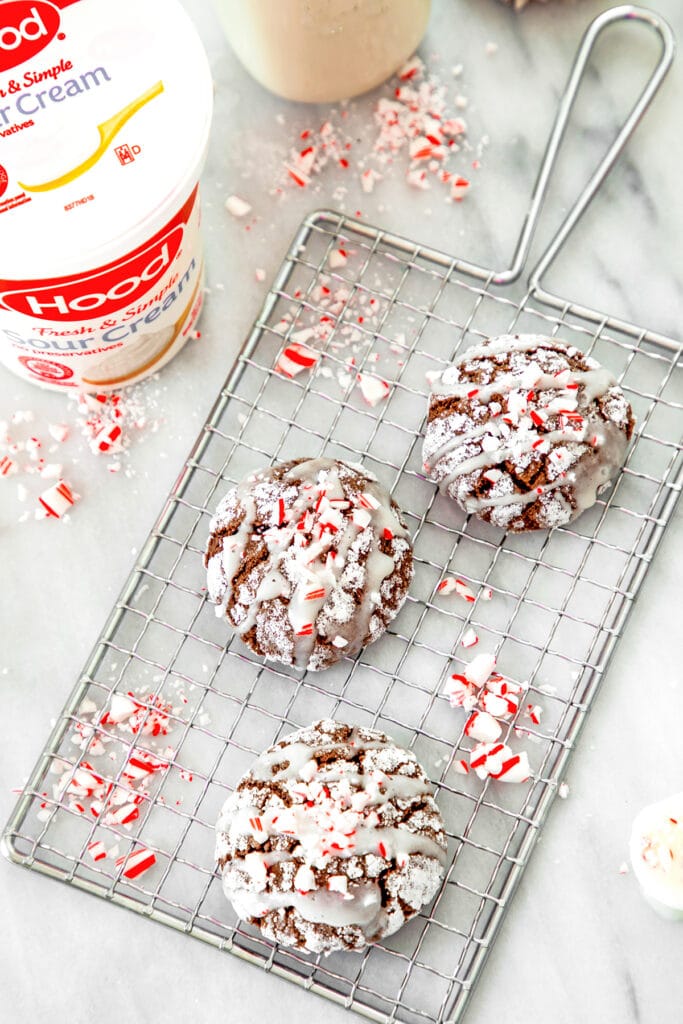 Bird's eye view of three chocolate peppermint sour cream crinkle cookies on a cooling rack on marble surface with crushed peppermint all around and sour cream container in background