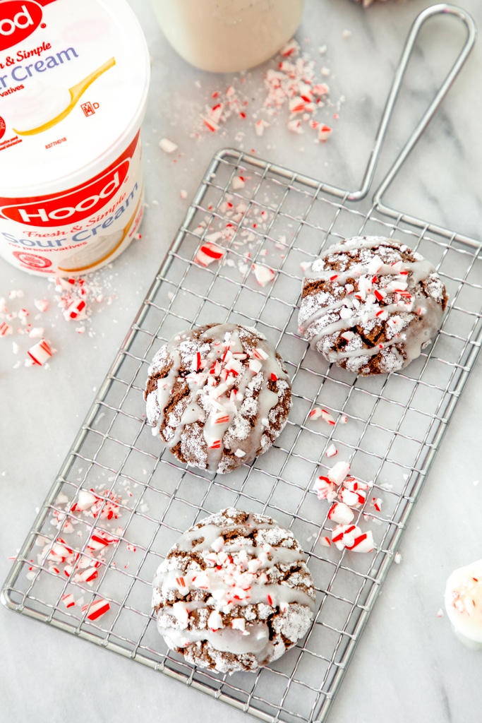 Bird's eye view of three chocolate peppermint sour cream crinkle cookies on a cooling rack on marble surface with crushed peppermint all around and sour cream container in background