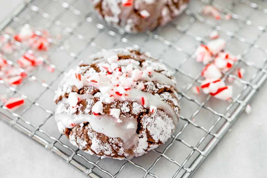 Landscape head-on closeup view of a chocolate peppermint sour cream crinkle cookie with icing and crushed candy canes on cooling rack