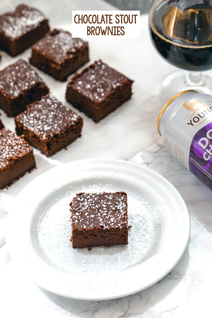Head-on view of a chocolate stout brownie on a white plate with more brownies, can of chocolate stout, and glass of chocolate stout in background with recipe title at top of image
