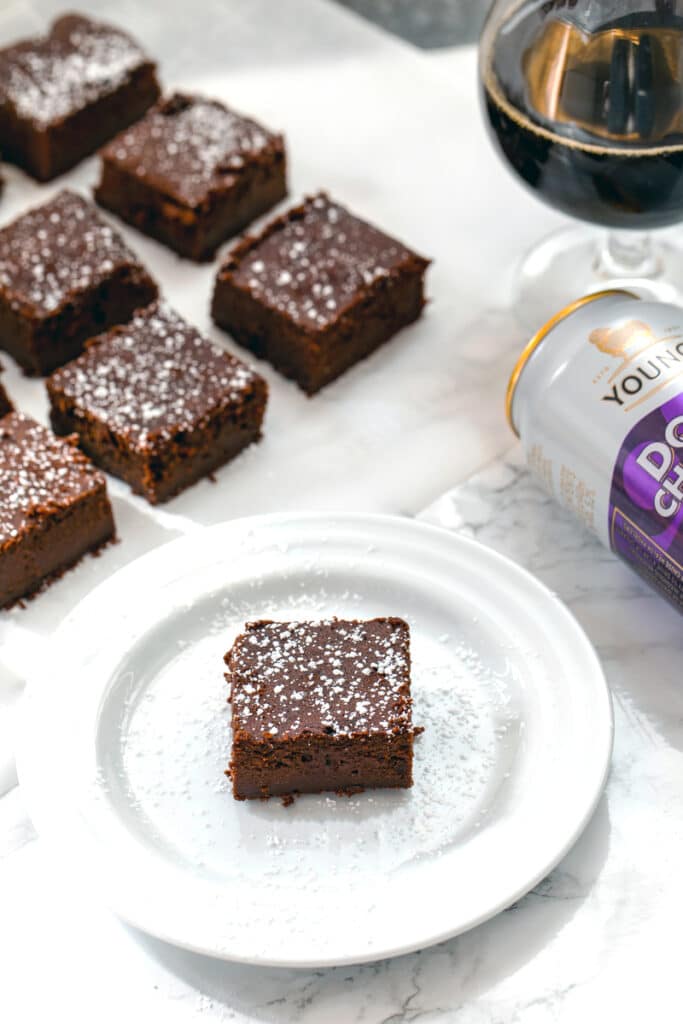 Head-on view of a chocolate stout brownie on a white plate with more brownies, can of chocolate stout, and glass of chocolate stout in background