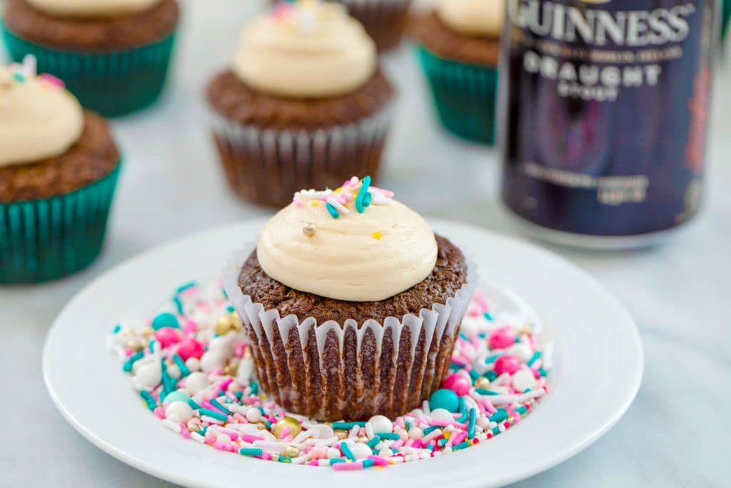 Landscape view of chocolate stout cupcake with Baileys frosting and sprinkles on a white plate covered in sprinkles with more cupcakes and can of Guinness in the background