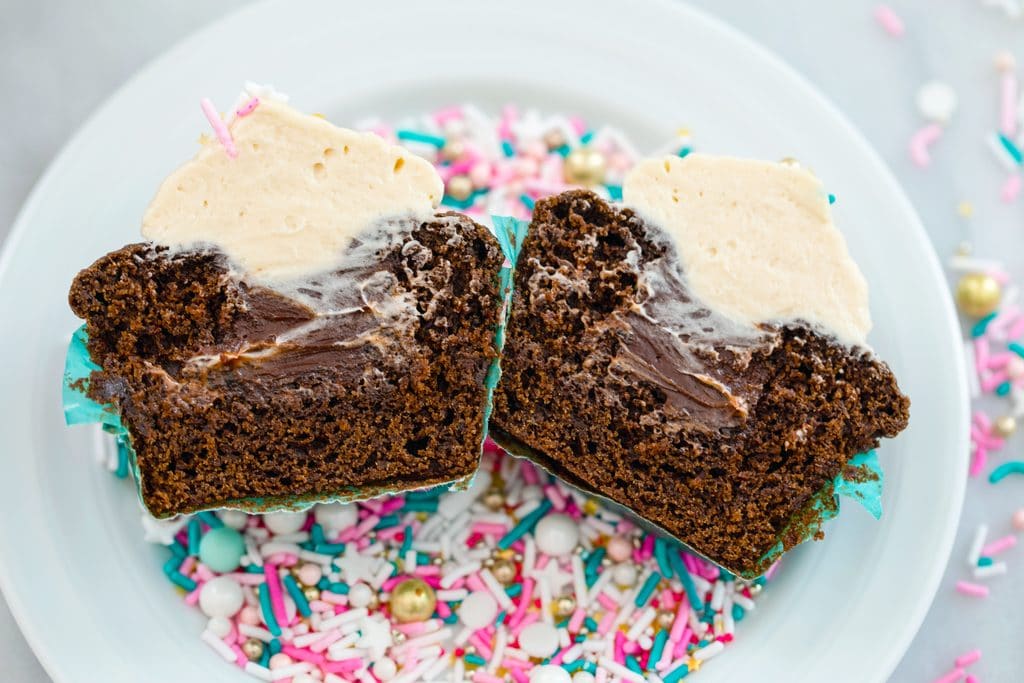 Overhead landscape view of a chocolate stout cupcake with Baileys frosting cut in half to show Irish whiskey filling inside sitting on a bed of sprinkles on a white plate