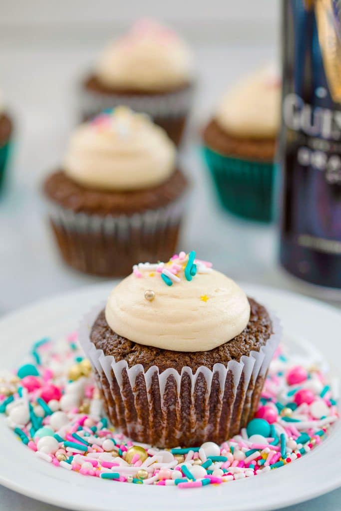 Head-on view of a chocolate stout cupcake with Baileys frosting and sprinkles on a white plate covered in sprinkles with more cupcakes and can of Guinness in the background