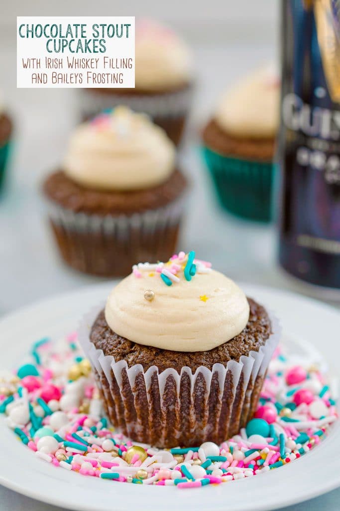 Head-on view of a chocolate stout cupcake with Baileys frosting and sprinkles on a white plate covered in sprinkles with more cupcakes and can of Guinness in the background and recipe title at top