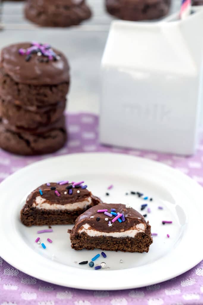 Overhead view of two chocolate surprise cookies cut open so you can see the marshmallow center with more cookies and ceramic milk carton in background