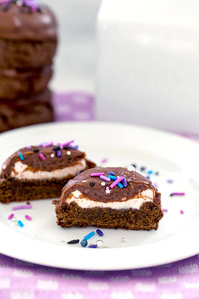 Head on view of chocolate surprise cookie cut in half to show marshmallow filling on white plate with sprinkles and stack of cookies in background