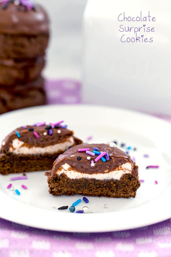 Head on view of chocolate surprise cookie cut in half to show marshmallow filling on white plate with sprinkles and stack of cookies in background with "Chocolate Surprise Cookies" text at top