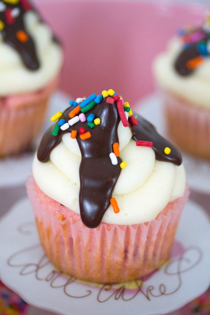 Head-on close-up view of a chocolate, vanilla, and strawberry cupcake with chocolate ganache drizzled on top and rainbow sprinkles.