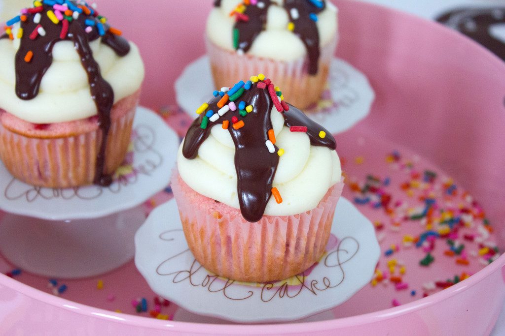 Landscape view of three chocolate, vanilla, and strawberry cupcakes all on little cupcake pedestals in a pink container with rainbow sprinkles all around