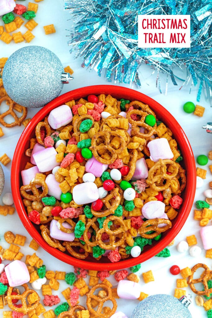 Overhead view of a bowl of Christmas trail mix with trail mix and ornaments on the table and recipe title at top