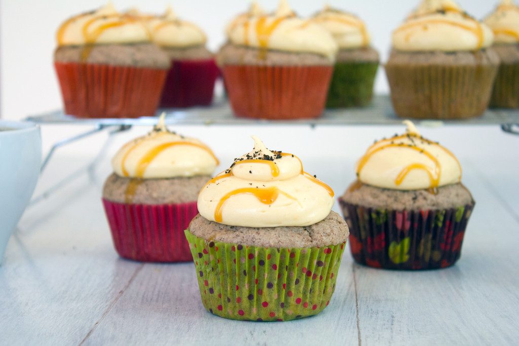 Landscape head-on view of three cinnamon cupcakes with pumpkin coffee cream cheese frosting with a baking rack with lots more cupcakes in the background