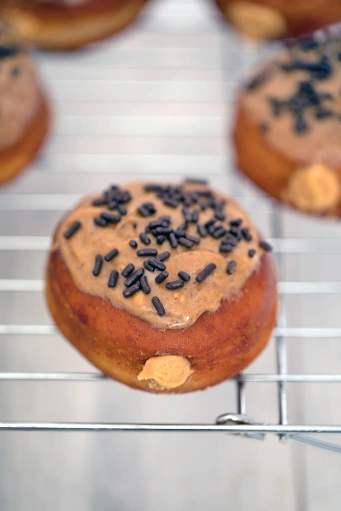 Overhead closeup view of a cinnamon doughnut topped with glaze and chocolate sprinkles with peanut butter cream filling peeking out on a baking rack.